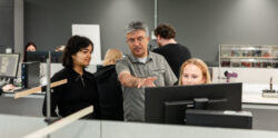 A lab with one person sat at a computer while two other people look at the screen, as the middle person of the three points at the screen.